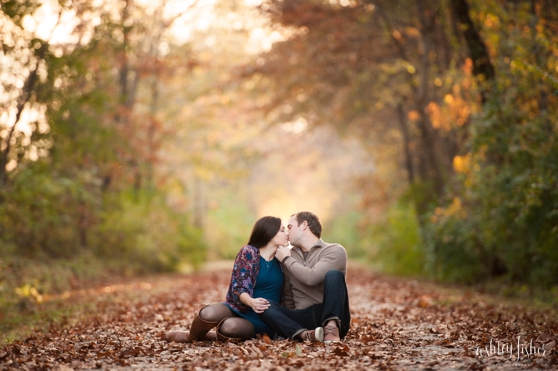 Photo of couple sitting and kissing on fall leaf covered trail